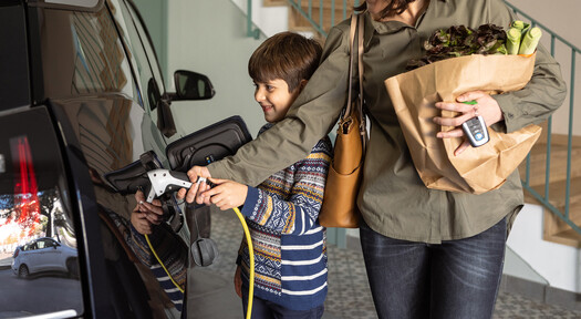 Eine Mutter und ihr 8-jähriger Sohn schließen zuhause nach dem Einkauf gemeinsam den Ladestecker ihrer Wallbox ans E-Auto an, um es zu laden.